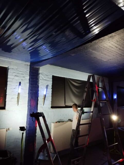 Lichtinstallation an schwarzer Decke in einem Raum mit Leiter und wei&szlig;er Wand.