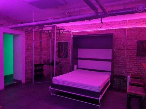 Beheiztes Schlafzimmer mit BDSM-Setup, Bett und Domszene, Pinkes Neonlicht, edle Backsteinwand.