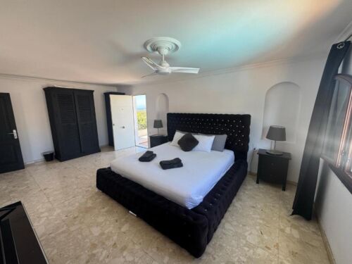 Ein modernes Schlafzimmer mit einem großen, schwarzen Bettgestell und weißen Bettwäsche, umgeben von Nachttischen mit Lampen, in einem hellen Raum mit weißen Wänden und großen Fenstern.