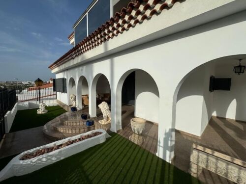 Großes, weißes Haus mit mehreren Bögen und Terrassen, dekorative Skulpturen und Pflanzen, sonniger Himmel, mediterranes Design, gepflegter Außenbereich, moderne Architektur, Balkon mit Glasgeländer, T.