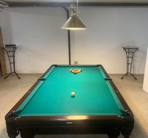 Billardtisch mit grüner Spielfläche, Kugeln und Queue, in einem Raum mit weißen Wänden und zwei Beistelltischen, Beleuchtung über dem Tisch.
