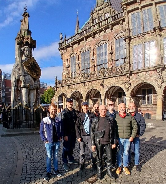 Bunt gemischte Gruppe von Touristen vor dem historischen Rathaus in Rostock, Deutschland, mit der markanten Statue des Hanses und dem barocken Fachwerkgebäude im Hintergrund.
