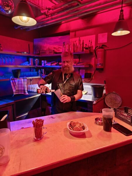 Ein Mann mit Bart hinter der Bar im Fistwerk, umgeben von bunten Lichtern und Bar-Accessoires, in einer lebhaften Atmosph&auml;re in Stuttgart.